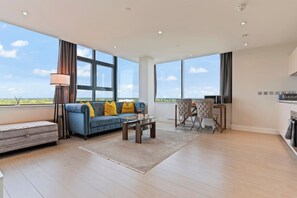 A spacious living room with a blue sofa, a coffee table, and a dining area with chairs. Large windows provide a view of the outdoors. - Heathrow Thorpe park Luxury Family Apartment - TURBO Wi-Fi (Sunbury-on-Thames)