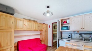 A compact kitchen with wooden cabinets, a microwave, and a small refrigerator. A red sofa is placed against the wall, and a wooden dining table with chairs is visible.
