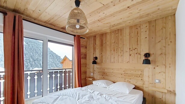 A bedroom with a wooden ceiling, a bed with white bedding, a window with curtains, and a view of snow-covered mountains. - VERONICA VR51 NATURE & MOUNTAIN 5 Pers (Châtel)