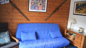 A room with a blue sofa, wooden walls, a wooden chair, a small wooden table with a lamp, and a framed picture on the wall.