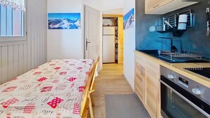 Een kleine keuken met houten kastjes, een ingebouwde oven en een magnetron. Er is een eethoek met een tafel en stoelen, en een raam met gordijnen.