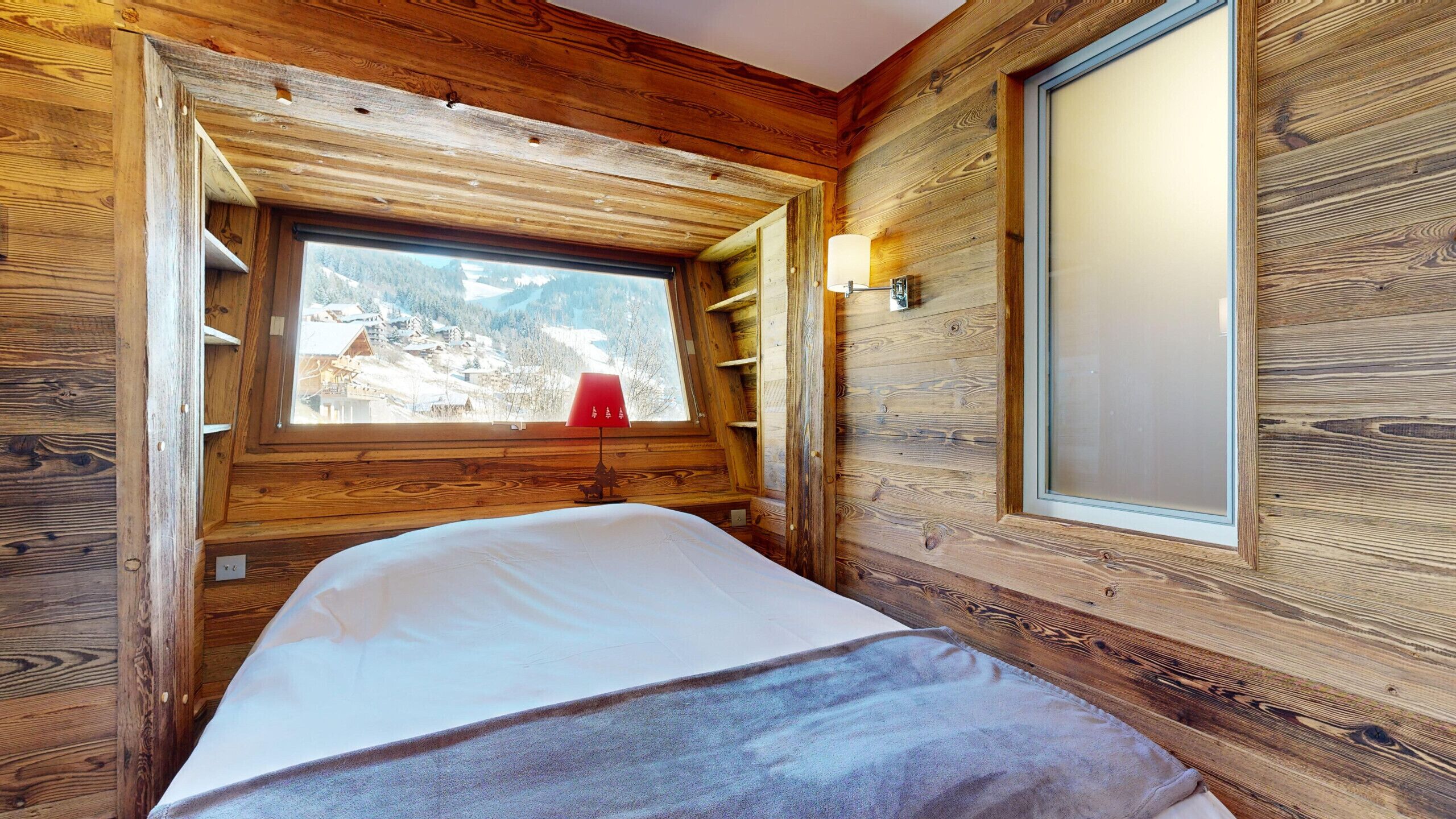 A wooden cabin room with a bed, a window with a view of a snowy landscape, and a wall-mounted light fixture.