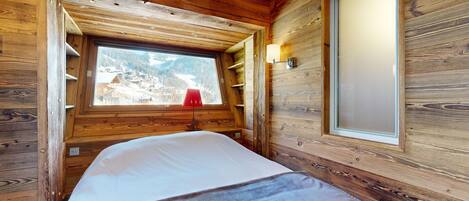 A wooden cabin room with a bed, a window with a view of a snowy landscape, and a wall-mounted light fixture.