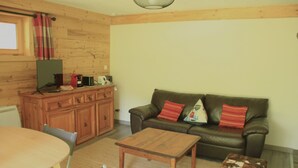 Un salon chaleureux avec un plancher en bois, un canapé en cuir marron agrémenté de coussins rouges, une table basse en bois, une table à manger avec des chaises, et un meuble en bois contenant une télévision et d’autres objets.