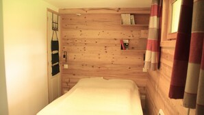 Une pièce étroite avec un mur en bois, un lit recouvert d’une couverture blanche, un radiateur et une étagère avec des livres et une petite boîte.