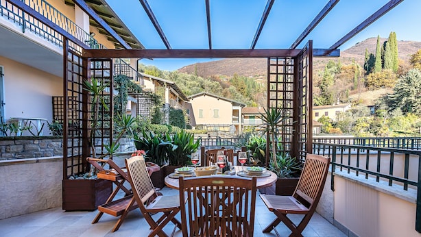 A covered outdoor dining area with wooden chairs and tables, surrounded by plants and overlooking a hillside with buildings. - Al Vittoriale 3 by Wonderful Italy (Gardone Riviera)