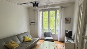 Un salon avec un canapé gris, une table basse blanche et un tapis à motifs. On y trouve une télévision posée sur une petite table, ainsi qu’une chaise recouverte d’un tissu gris. Une grande fenêtre ornée de rideaux blancs laisse entrer la lumière naturelle dans la pièce.