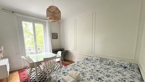 Une chambre à coucher comprenant un lit, une table, des chaises, un luminaire suspendu et une fenêtre avec des rideaux.