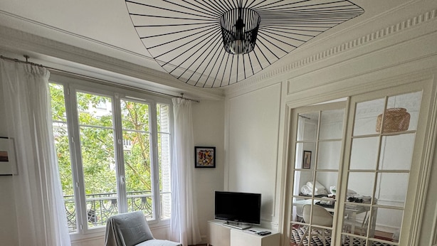 Un salon moderne avec un canapé, une table basse et un téléviseur. Une grande fenêtre, ornée de rideaux, offre une vue sur la verdure environnante.