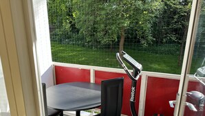 Ein Balkon mit Tisch und Stühlen, einem Reebok-Hometrainer und Blick auf einen grünen Garten durch ein Drahtgeflecht.
