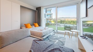 Ein modernes Schlafzimmer mit Bett, Sofa, Schreibtisch und einem großen Fenster mit Blick auf ein Gebäude und einen Garten.