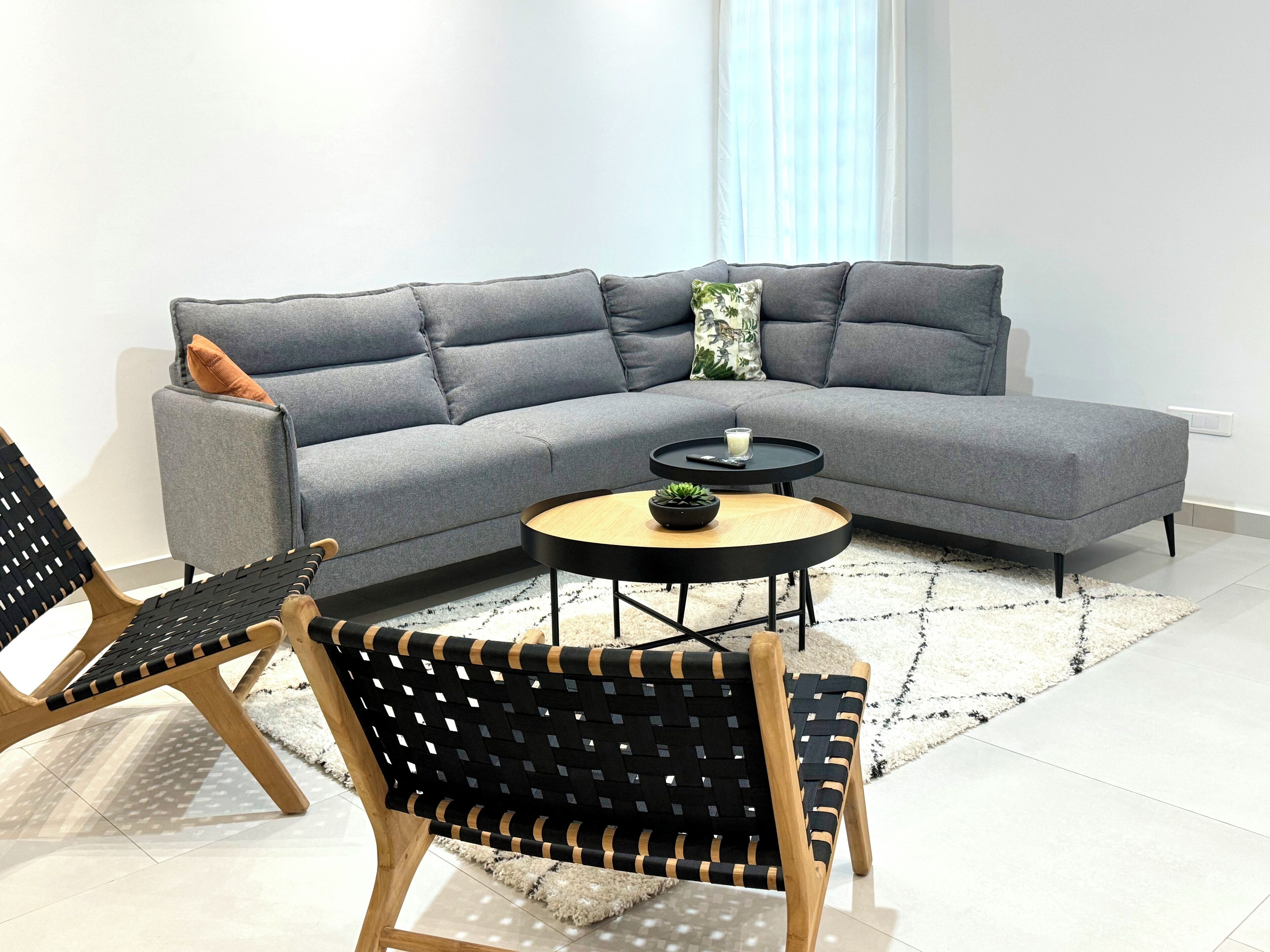 Una sala moderna con un sofá seccional gris, una mesa redonda de centro y dos sillas tejidas de madera y color negro.