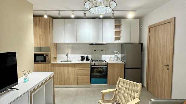 A modern kitchen with white cabinets, a built-in oven, and a microwave. There is a wicker chair and a sofa in the living area. - KIA ORA APPART HOTEL (Abidjan)