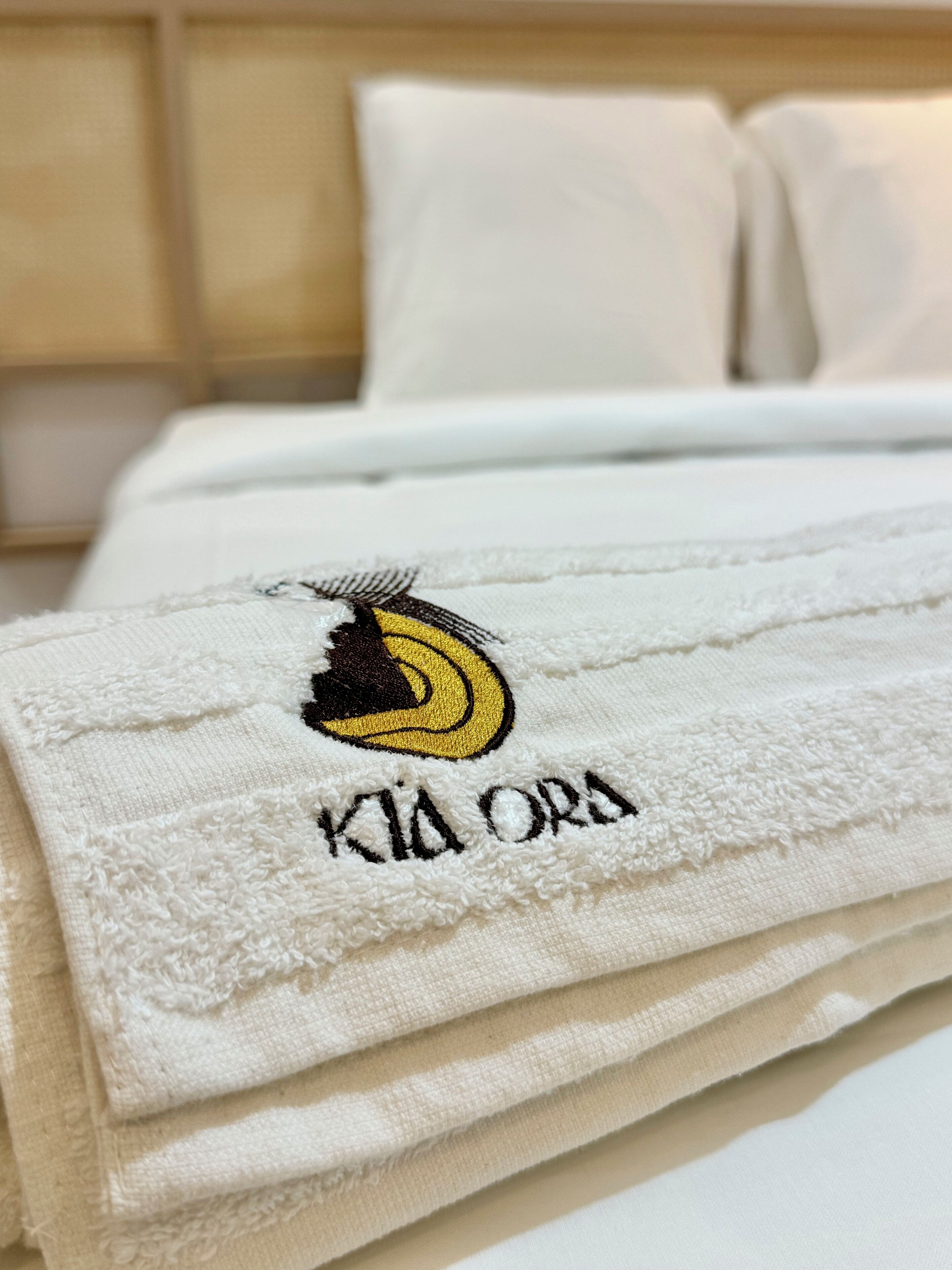 A neatly folded white towel with a logo and text on it, placed on a bed with white bedding and pillows.