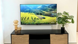 A flat-screen TV mounted on a wall above a wooden and black TV stand with a plant and two glasses on it. - KIA ORA APPART HOTEL (Abidjan)