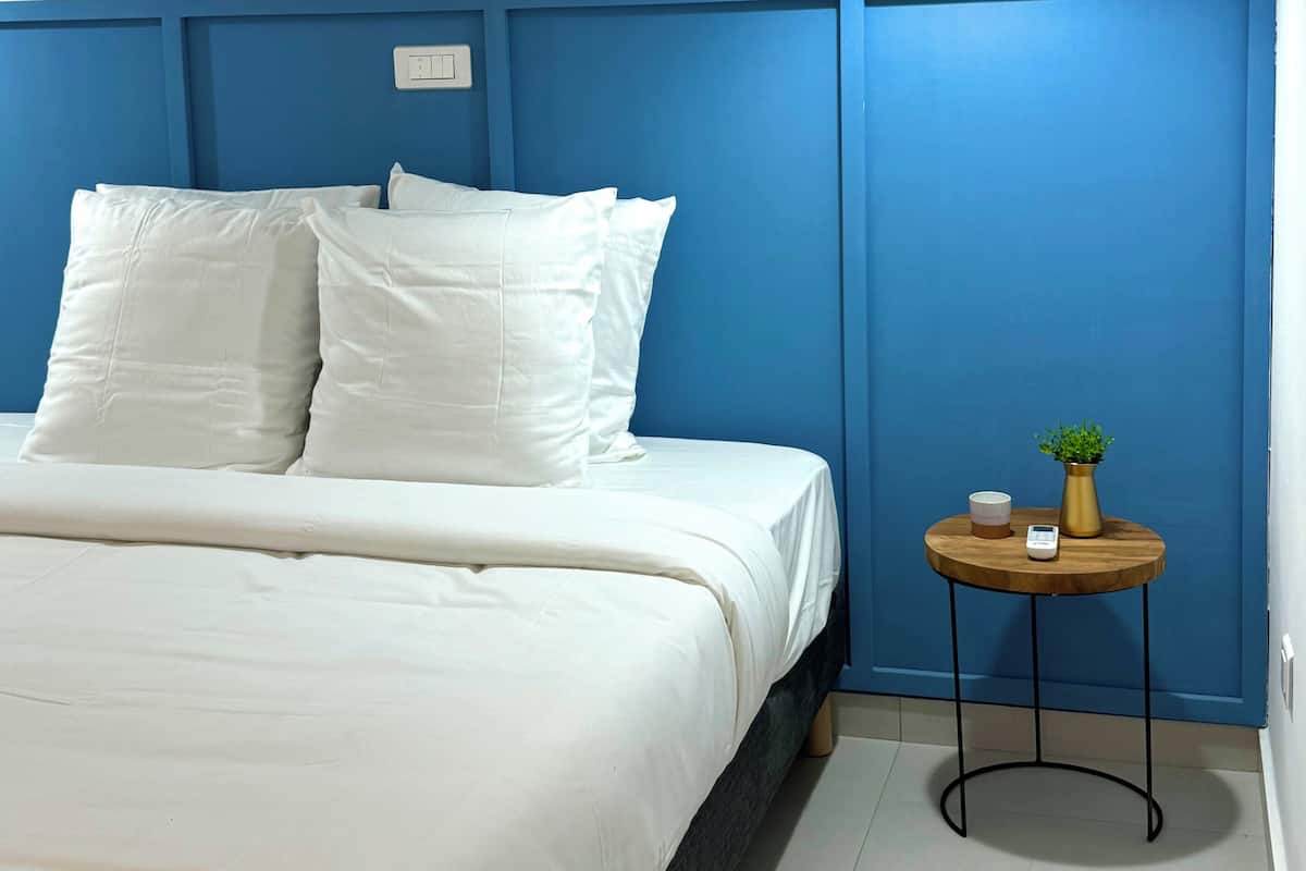 A bedroom with a bed, white bedding, a bedside table with a plant, and a wall-mounted light fixture.