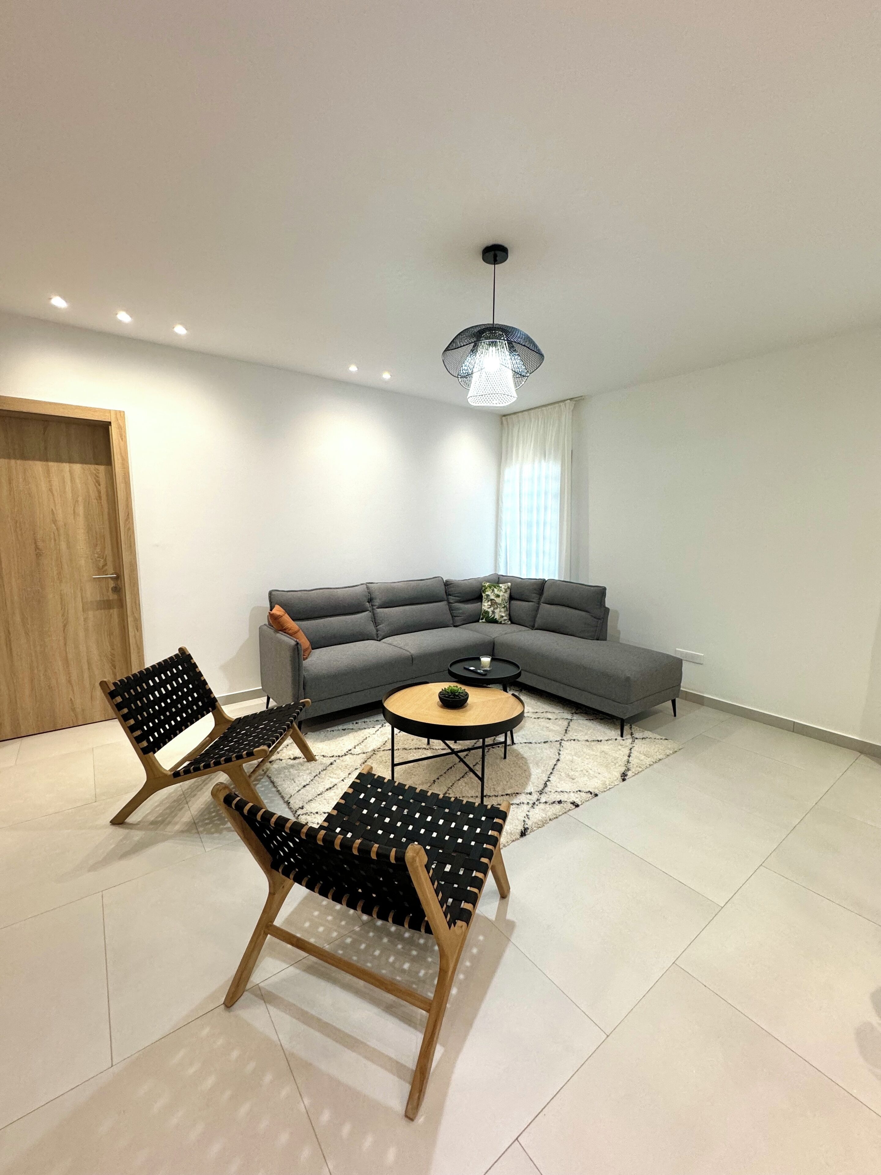 Un salon moderne avec un canapé d’angle gris, deux chaises noires en bois, une table basse ronde en bois et un luminaire suspendu.