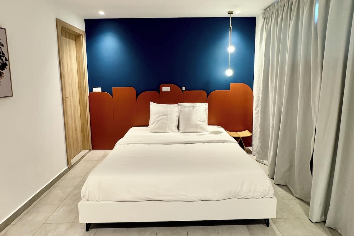 A modern hotel room with a large bed, a dark blue accent wall, and a large window with curtains.