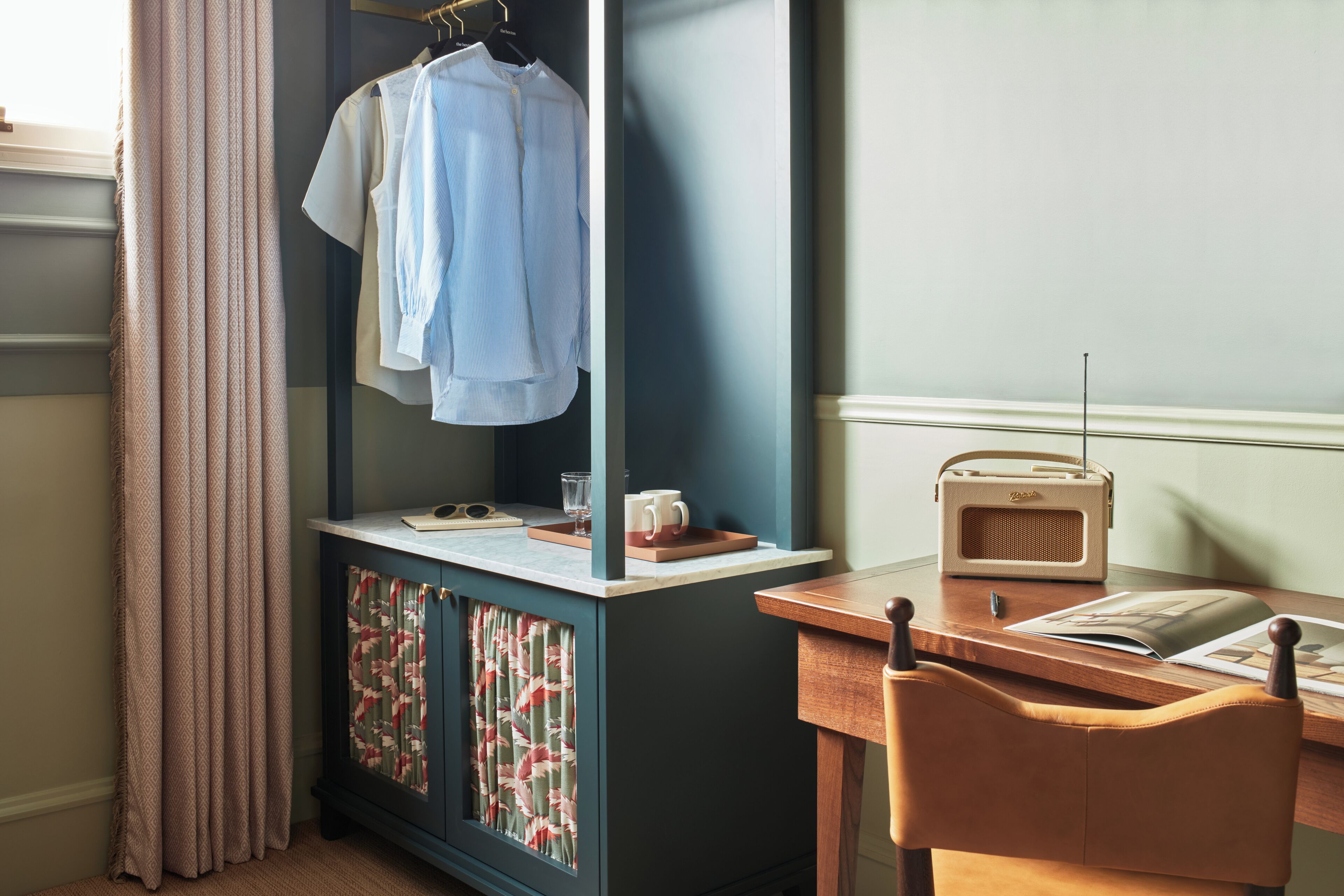 A room with a desk, a chair, a radio, and a wardrobe with hanging clothes.