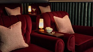Una habitación oscura con sofás de terciopelo rojo, una mesita con un cuenco de palomitas de maíz y una lámpara encendida.