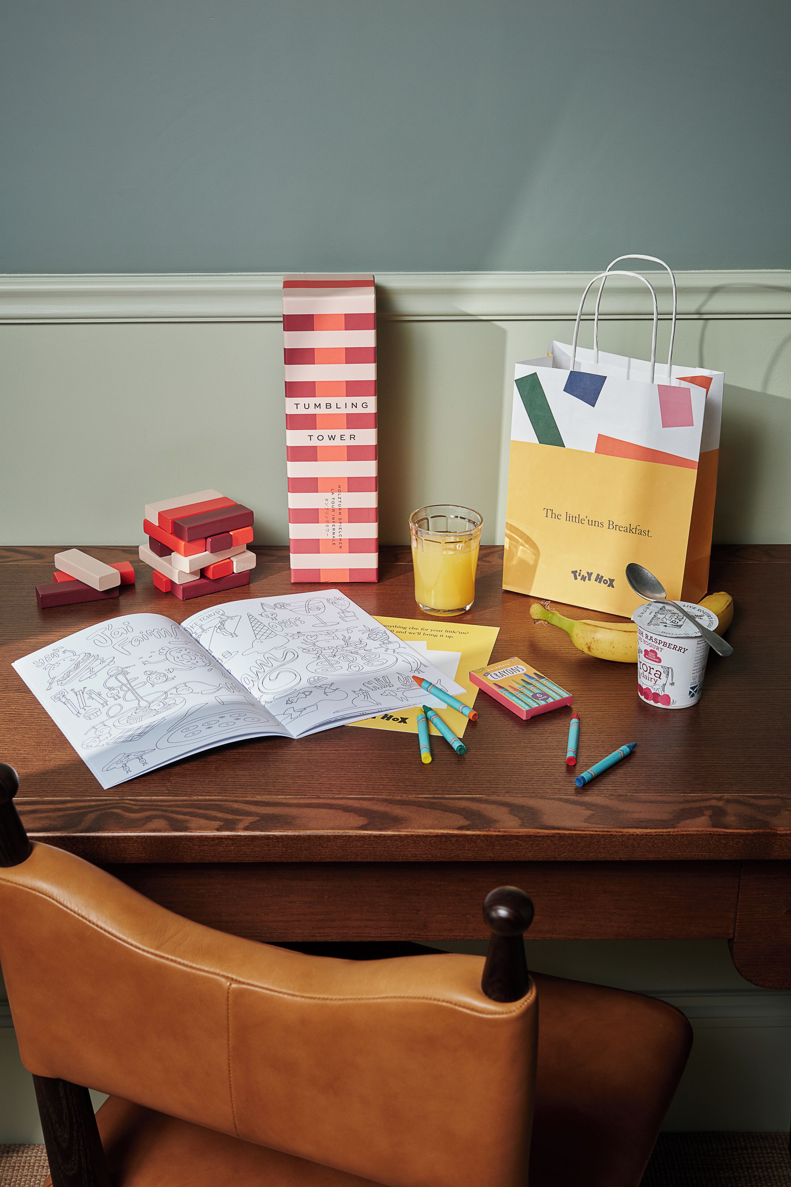 A wooden table with a book, a glass of juice, a bag of TUMBLING TOWER, and some stationery.