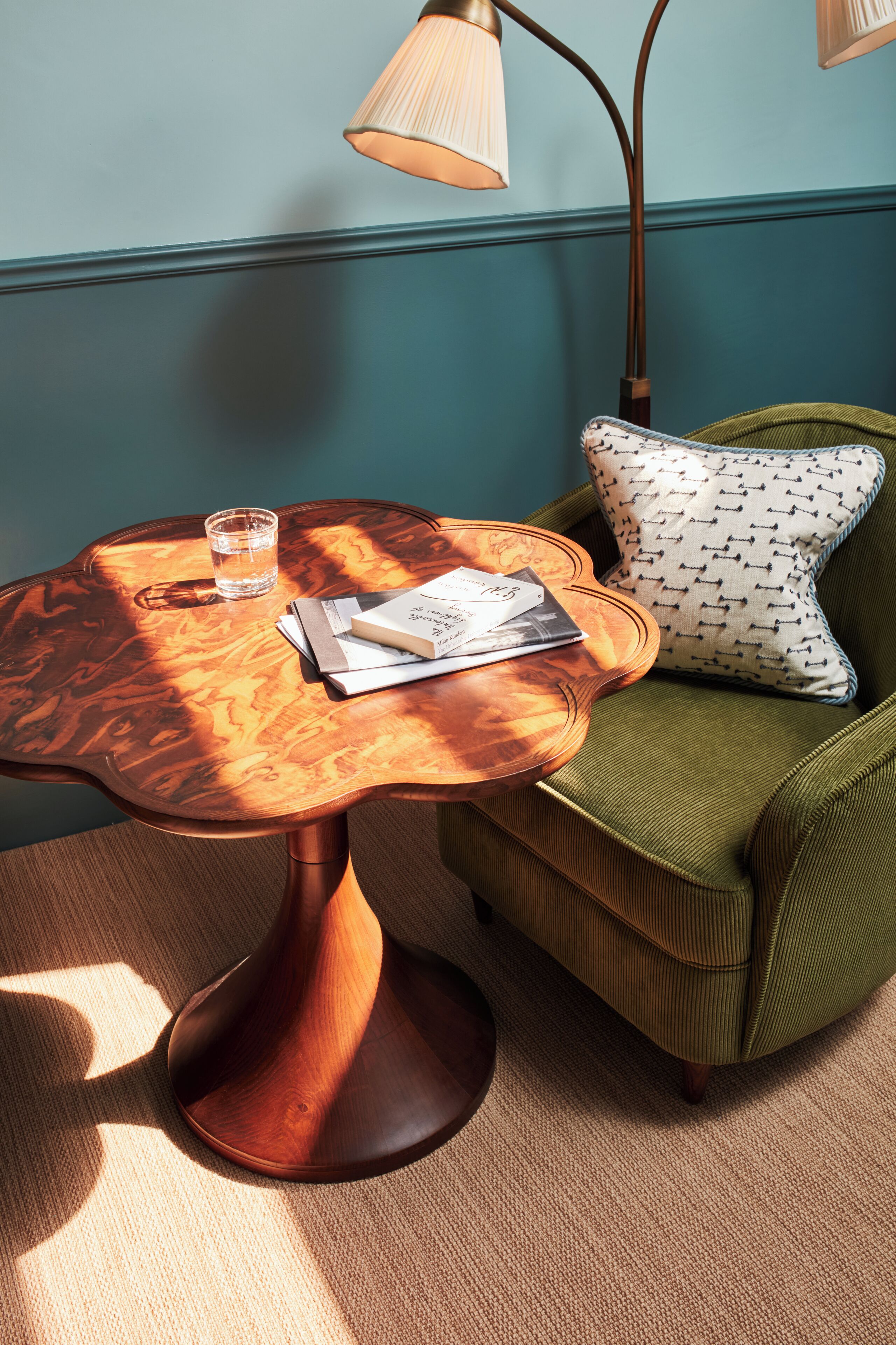 A room with a wooden table, a green armchair, a lamp, and a glass of water.