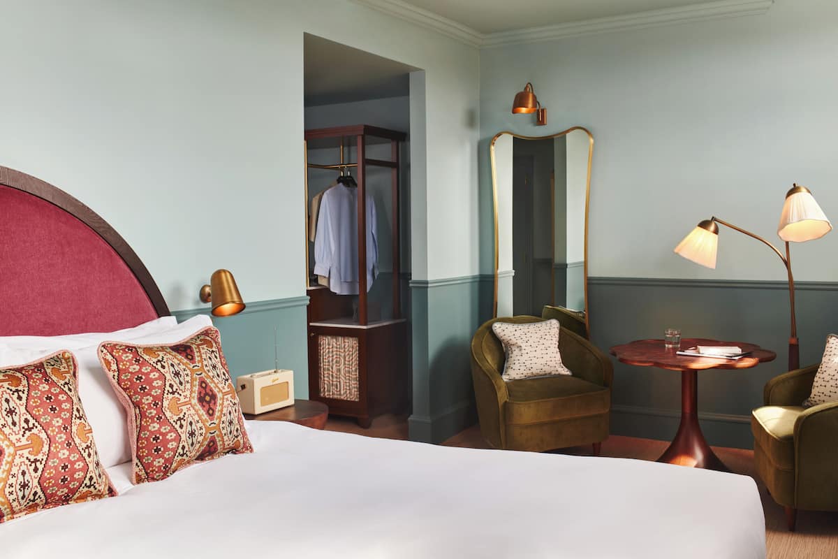 Ett hotellrum med en stor säng, två fåtöljer, ett litet bord, en spegel, en garderob och en lampa.