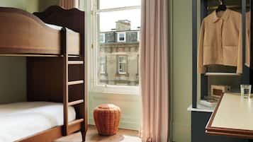 Una cama individual con cabecera de madera, un pequeño taburete redondo, una mesa con un vaso y un perchero.