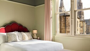 Una cama bien hecha con cabecera roja, una lámpara de noche y una ventana con vistas a edificios históricos.