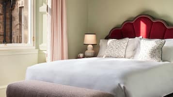 A neatly arranged bedroom with a large bed, a bedside table with a lamp, a window with curtains, and a patterned rug.