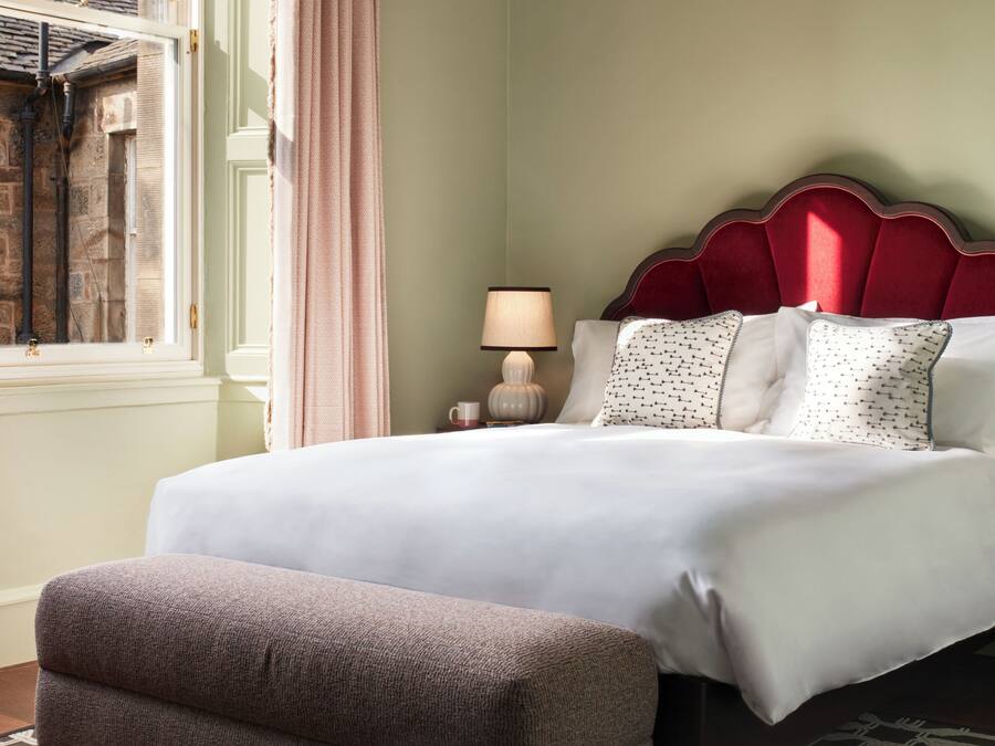 A neatly arranged bedroom with a large bed, a bedside table with a lamp, a window with curtains, and a patterned rug.