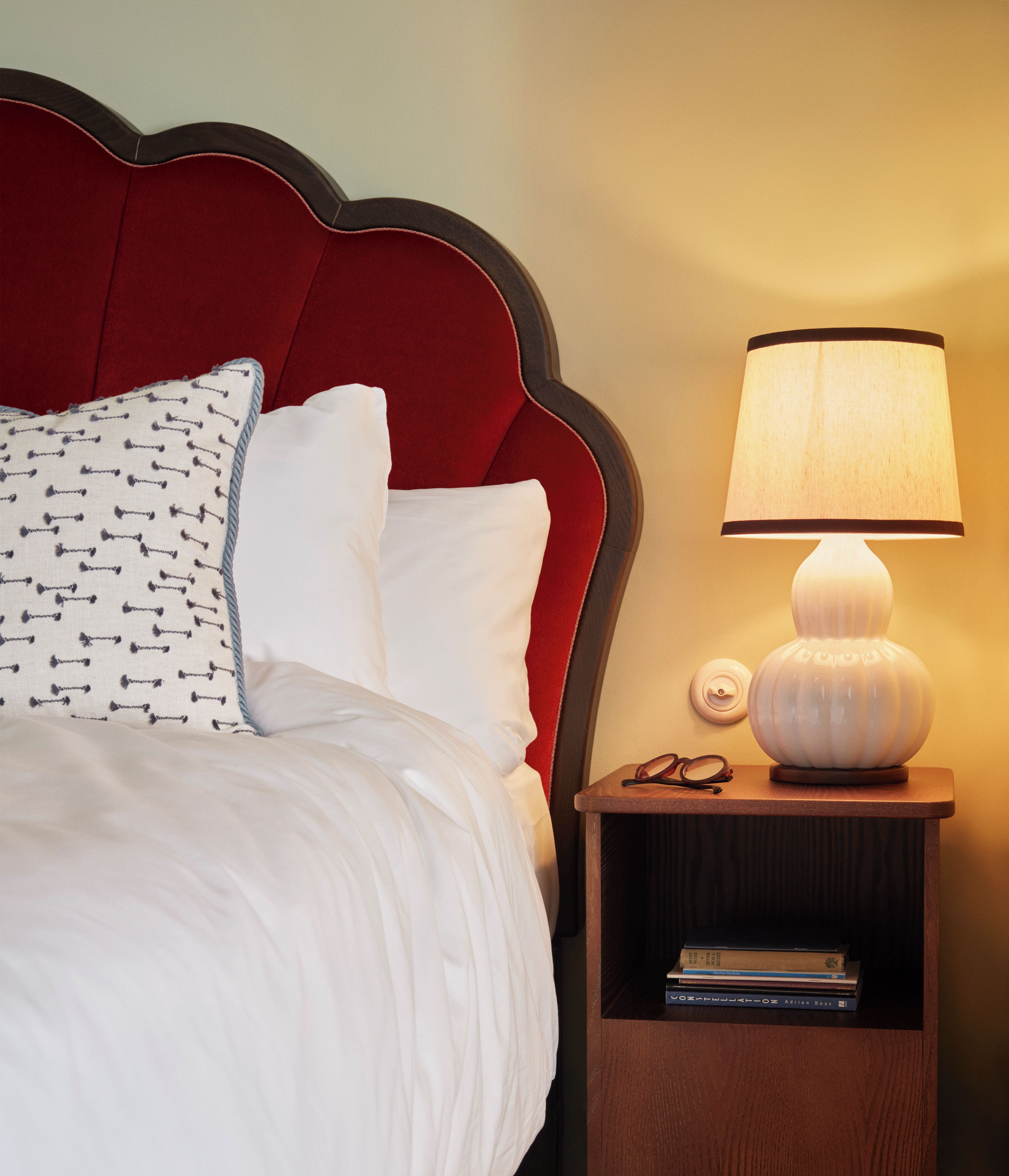 A neatly made bed with white linens and a red headboard, a bedside lamp, and a small wooden nightstand with books and glasses.