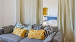 Una sala moderna con un sofá gris decorado con cojines estampados, vista a un dormitorio a través de una ventana con cortinas, y una pared de acento azul.