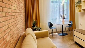 A modern living room with a brick wall, a beige sofa, a dining table with chairs, and a chandelier. - PoshSpot Apartments (Cluj-Napoca)