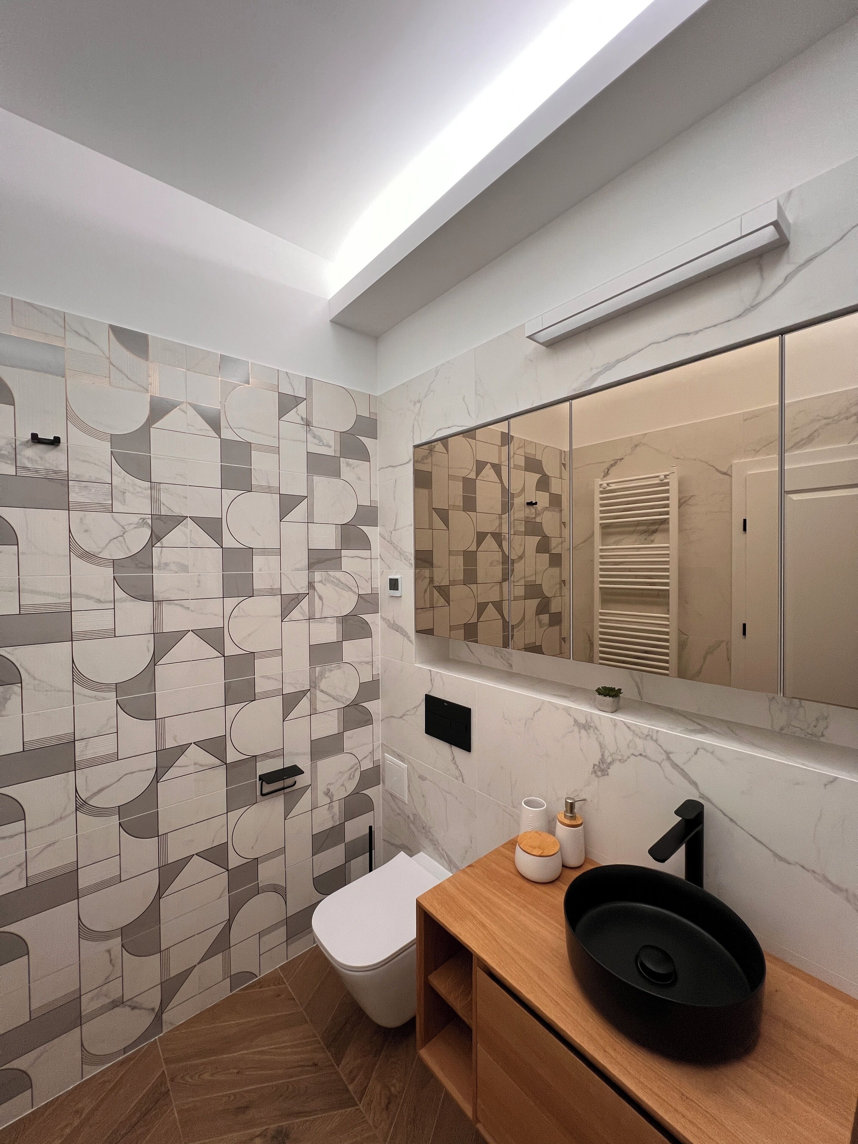 A modern bathroom with geometric patterned tiles, a white toilet, a wooden vanity with a black sink, and a mirror above the vanity.