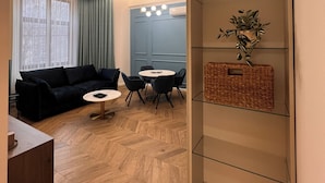 A modern living room with a sofa, a coffee table, and a dining area with chairs. There is a plant on a shelf and a window with curtains. - PoshSpot Apartments (Cluj-Napoca)