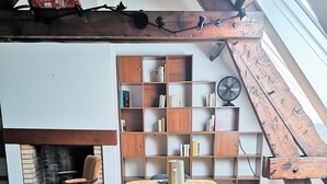Un coin repas avec une table en bois dressée pour quatre personnes, entourée de chaises noires. On aperçoit, à l’arrière-plan, une bibliothèque remplie de livres et un ventilateur.