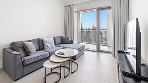 Una sala de estar moderna con un sofá gris, dos mesas redondas de centro, un televisor de pantalla plana y un amplio ventanal corredizo que da a un balcón con vistas a la ciudad.