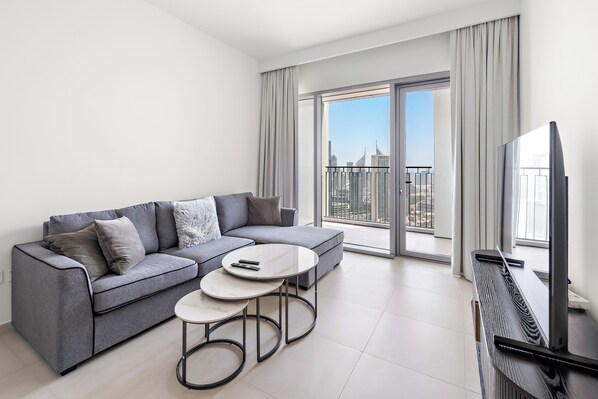 A modern living room with a grey sofa, two round coffee tables, a flat-screen TV, and a large sliding glass door leading to a balcony with a city view.