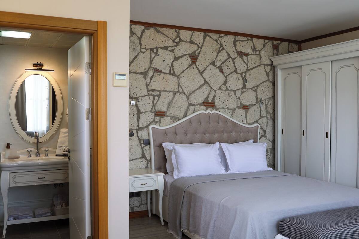 Un dormitorio con pared de piedra, una cama con ropa blanca, una mesita de noche, un tocador y un espejo.