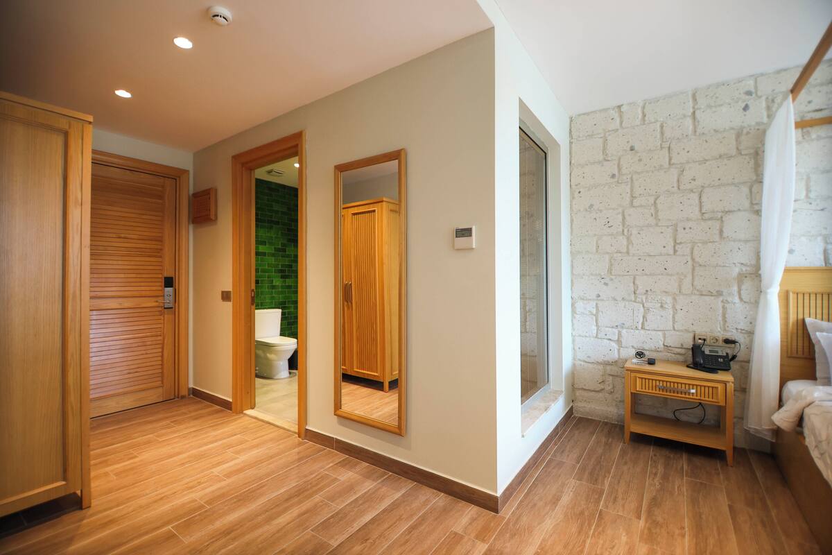 Habitación de hotel con suelo de madera, pared de ladrillo, puerta de madera, mesita de noche pequeña con radio y cama con cabecero blanco.
