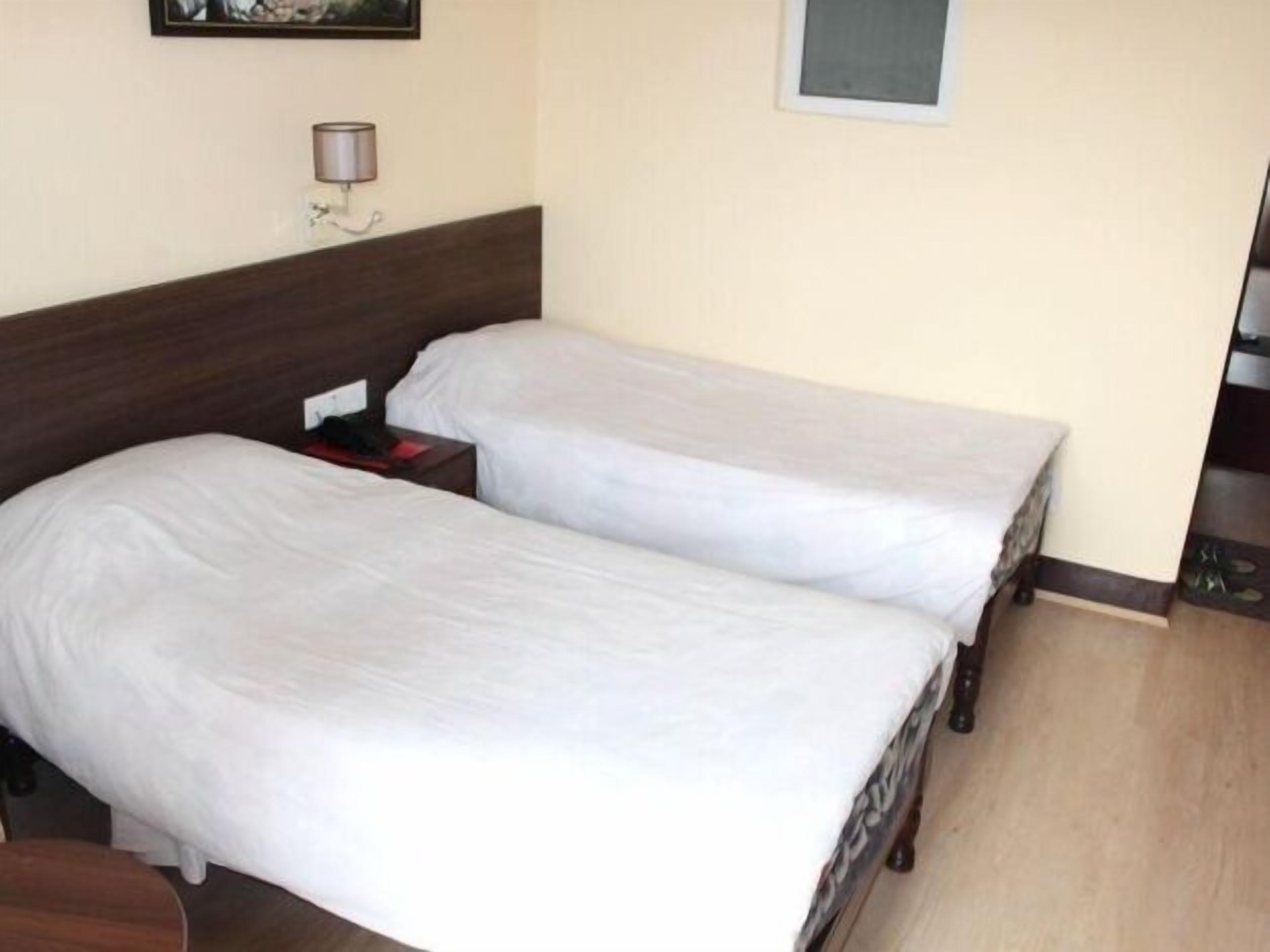 A hotel room with two single beds, a wooden headboard, a bedside table with a lamp, and a small window.