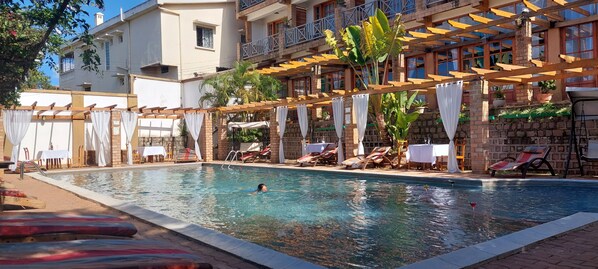 En swimmingpool med solstolar och parasoller, omgiven av en byggnad med balkonger och planterade blommor i krukor.