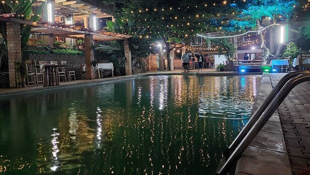 Zona de piscina con superficie de agua cristalina que refleja luces, rodeada por un área de estar cubierta con mesas y sillas, y un edificio con balcón.