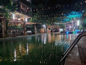 Zona de piscina amb superfície d'aigua nítida que reflecteix llums, envoltada per una zona coberta de seients amb taules i cadires, i un edifici amb balcó.
