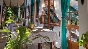 Un comedor con mesas dispuestas para dos, ventanas amplias con cortinas y plantas en macetas.