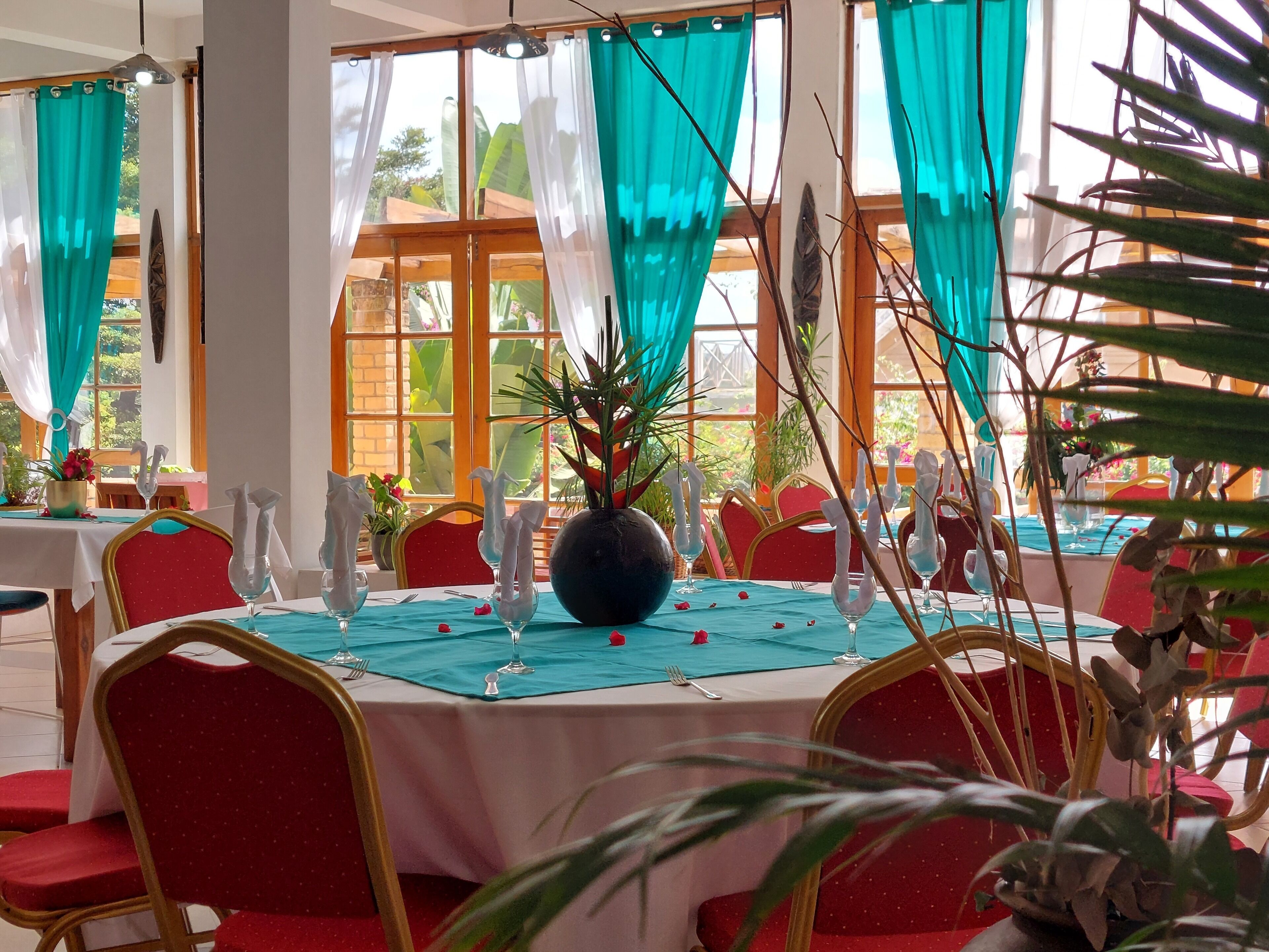 Comedor con mesas dispuestas para una comida, con manteles color turquesa, sillas rojas y un jarrón central con flores.