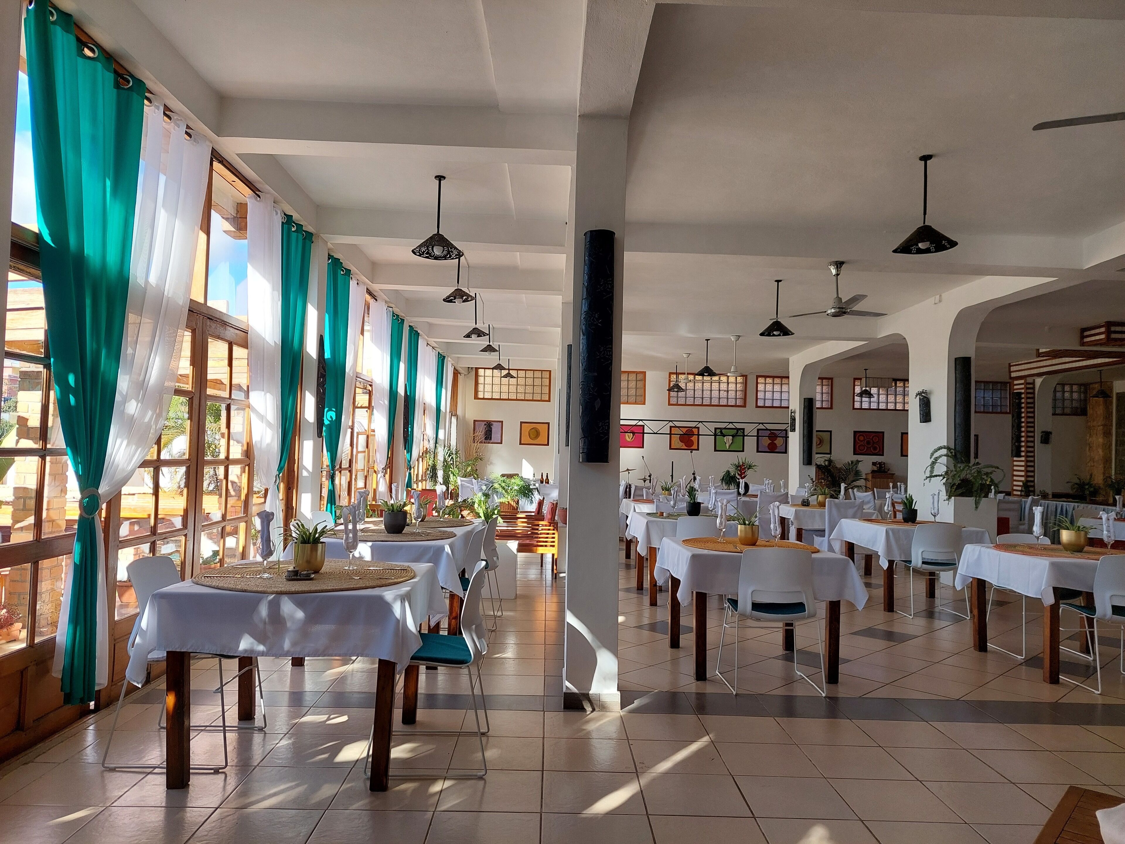 Un restaurant spacieux, doté de nombreuses tables et chaises, de grandes fenêtres avec des rideaux et de ventilateurs de plafond.