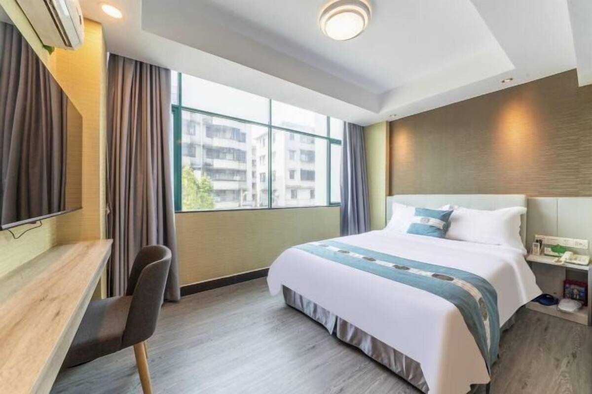 Une chambre d’hôtel avec un lit, un bureau et une chaise. Il y a une fenêtre avec des rideaux, un climatiseur fixé au mur et une table de chevet avec une lampe.