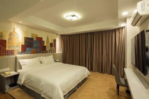 Una habitación de hotel con cama, escritorio, silla, un cuadro de un paisaje urbano y un equipo de aire acondicionado.